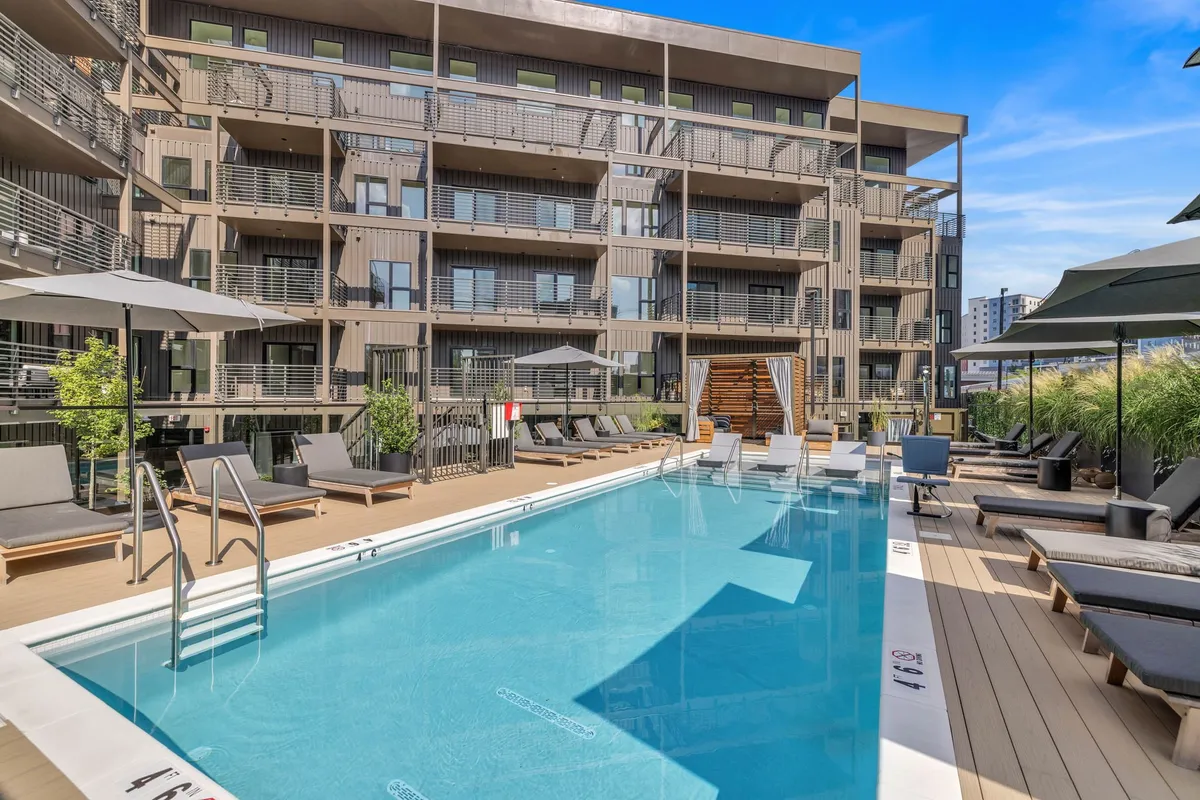 A vibrant outdoor swimming pool set against a modern apartment building, featuring ample lounge seating and inviting spaces for guests to swim, sunbathe, or socialize. The pool is surrounded by greenery and offers a resort-style atmosphere.