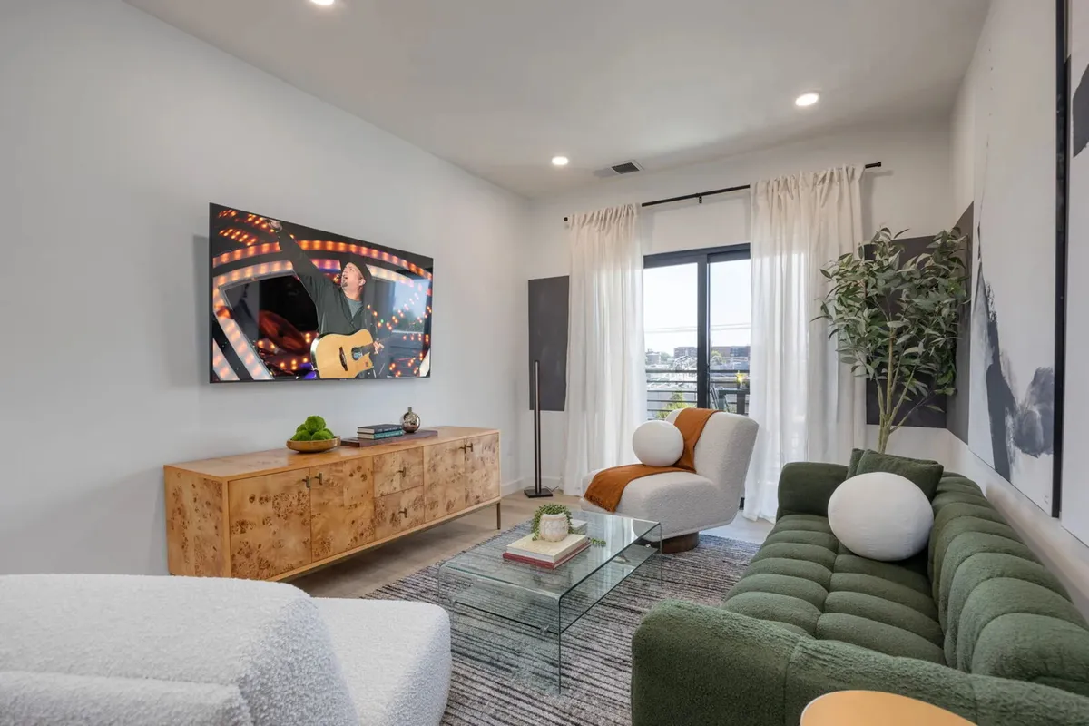 A spacious living area with a green tufted sofa, large TV ready for movie nights, a glass coffee table, and ample natural light from a picture window. The room is designed for relaxation and social gatherings, with vibrant décor and comfortable furnishings that invite conversation and entertainment.