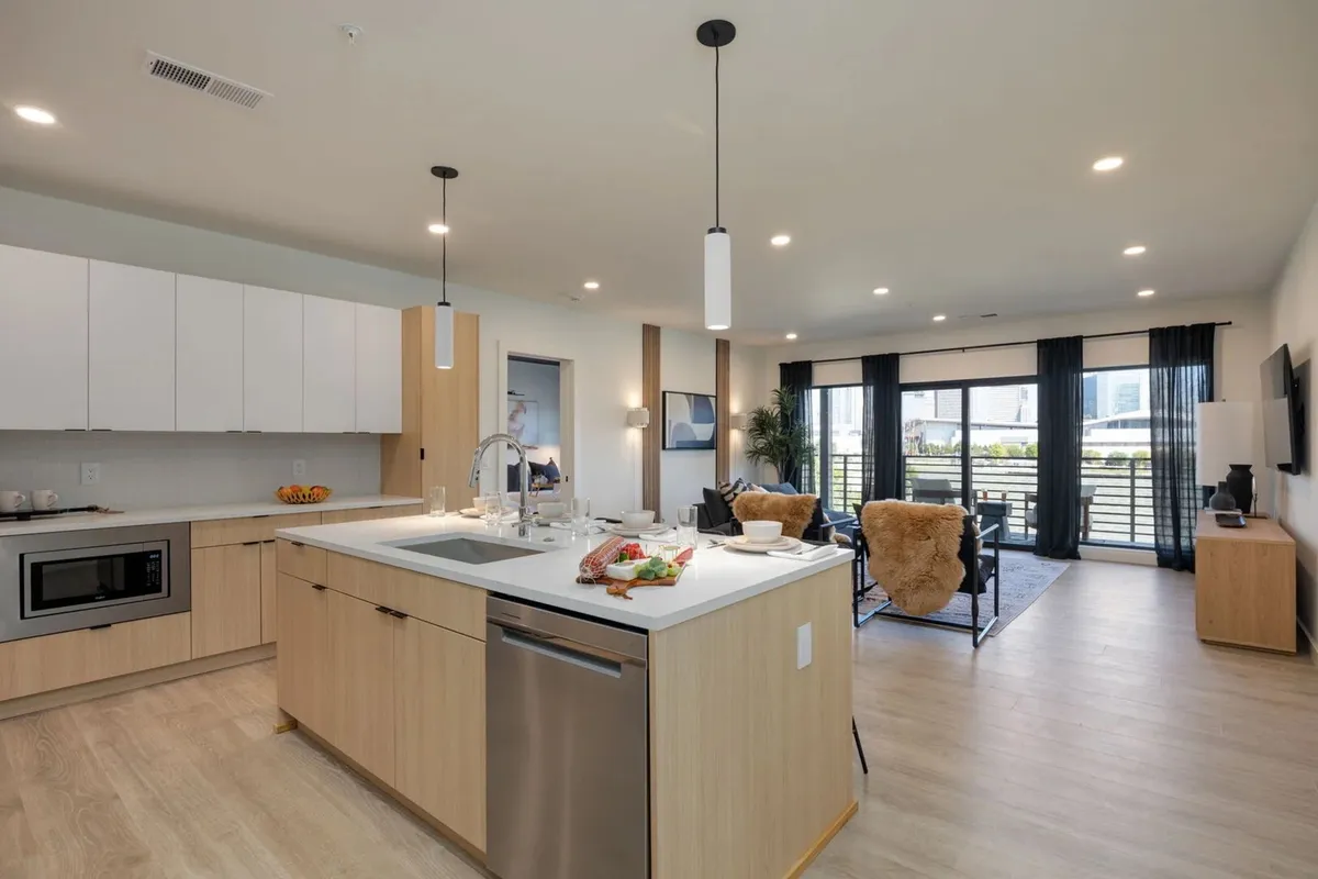 A stylish kitchen island anchors the scene, with modern appliances and inviting living area seating nearby. Large windows usher in daylight, while decorative plants and contemporary accents create a welcoming, cozy environment ideal for group activities like dining or game nights.