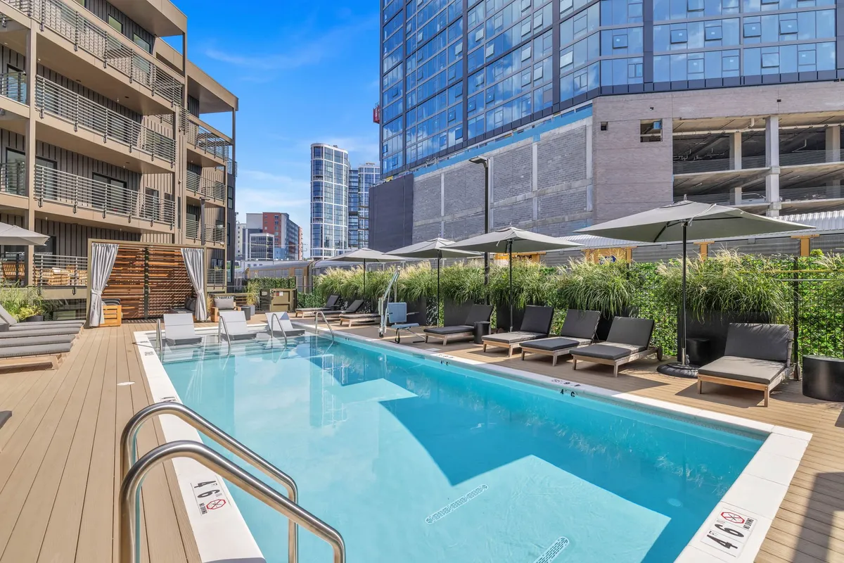 A contemporary outdoor pool area with sparkling blue water, surrounded by modern lounge chairs, umbrellas, lush greenery, and sleek buildings in the background. The setting is perfect for sunbathing, socializing, or relaxing with a drink by the water.