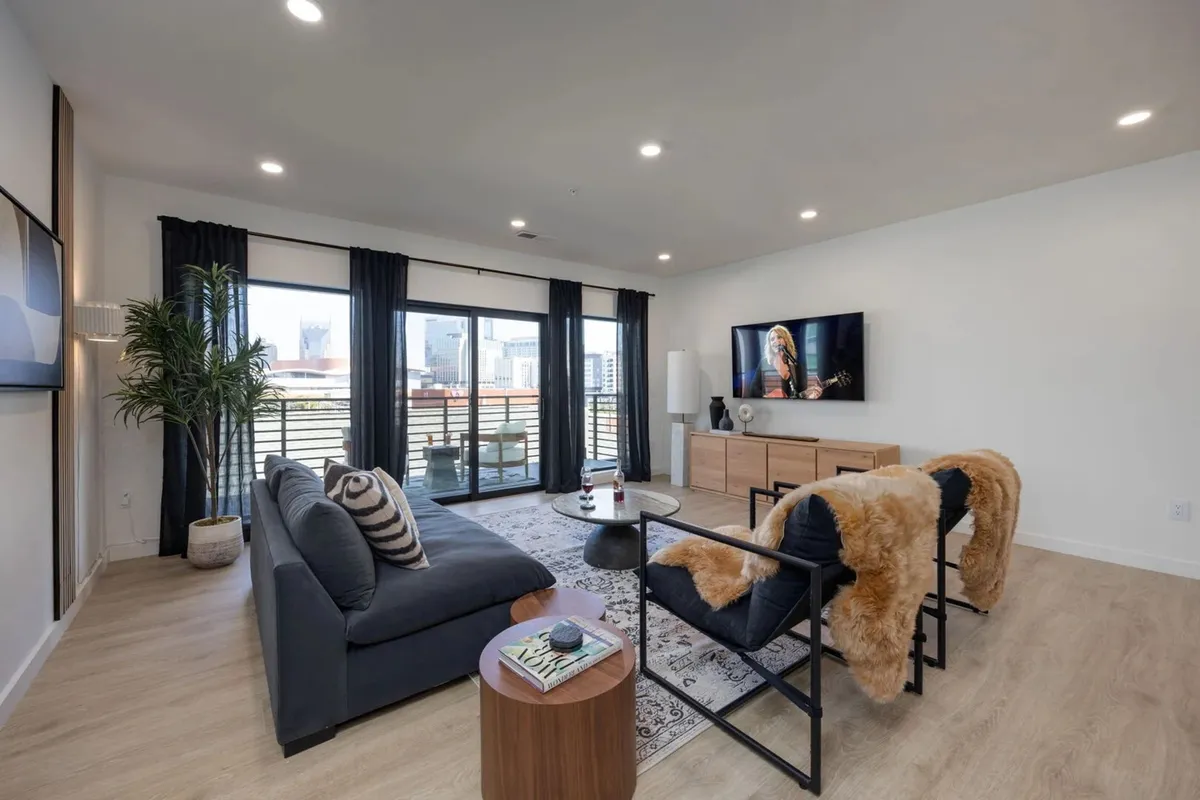 The image showcases a spacious, stylish living area with a plush sofa, cozy armchairs, and a sleek coffee table arranged for relaxation and conversation. Large windows frame vibrant city skyline views, filling the room with natural light. A mounted television provides entertainment, while the chic decor creates a welcoming, contemporary atmosphere perfect for gathering with friends or family.