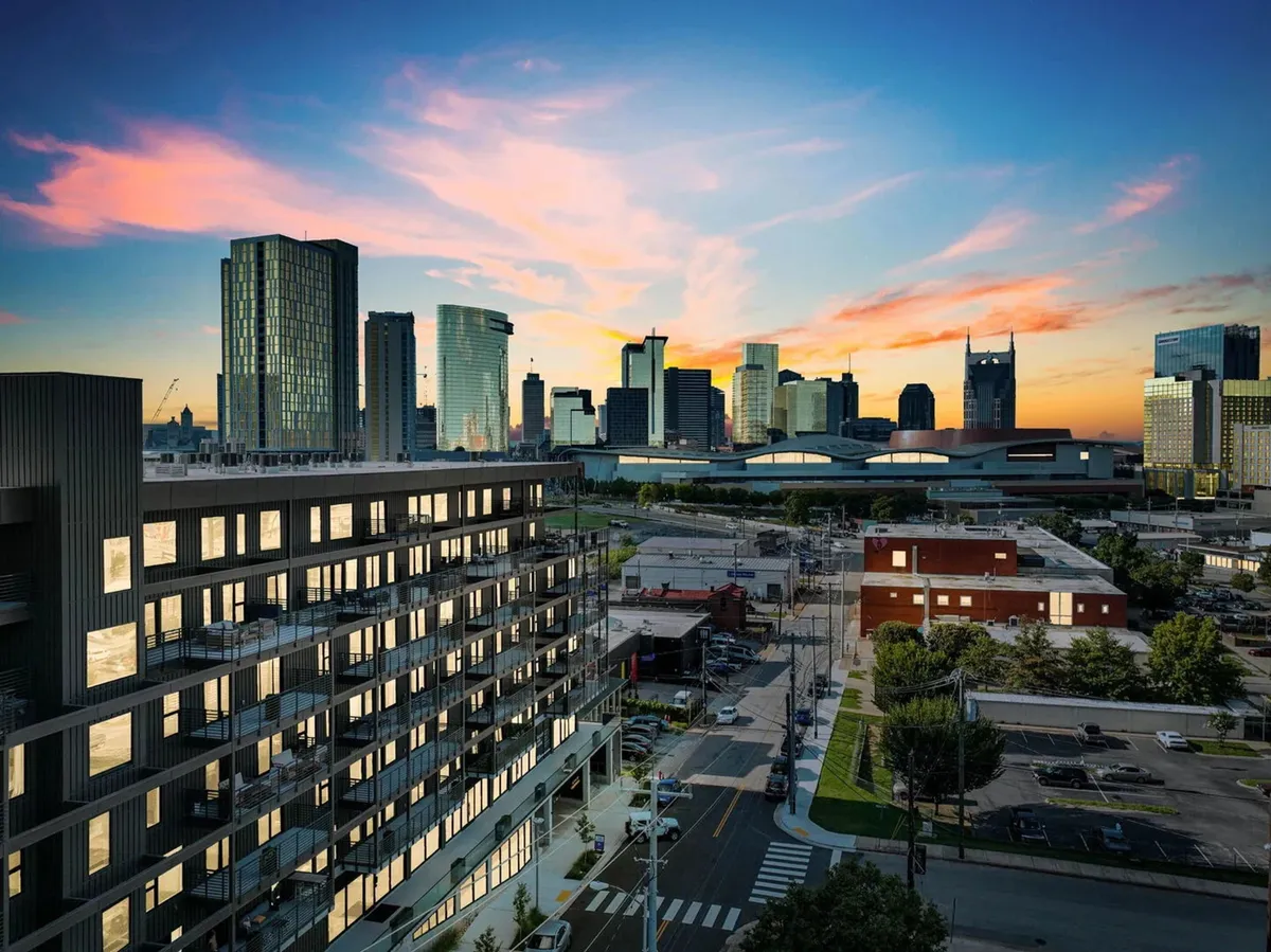 A vibrant exterior view showcasing the modern architecture of a residential building against the Nashville city skyline at sunset. The scene includes an open street, colorful evening sky, and hints of the urban energy surrounding the property.