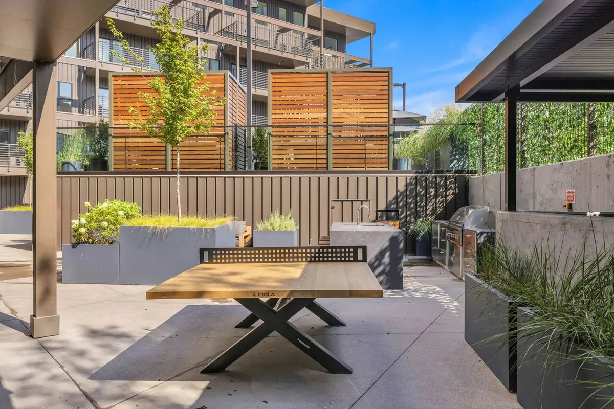 An outdoor communal area with a wooden table, decorative screens, green plants, and a modern grill setup. The relaxed atmosphere is ideal for barbecues, group meals, and evening get-togethers.