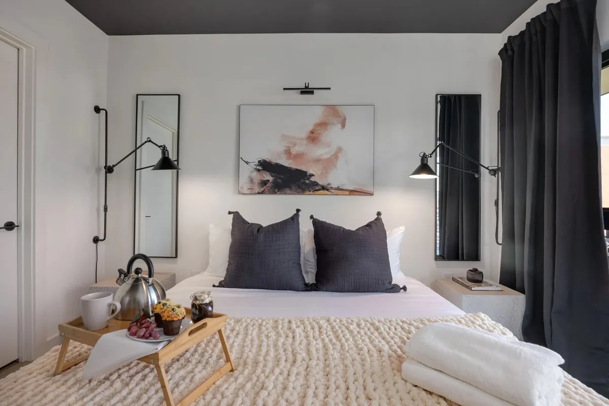 A serene bedroom featuring a plush bed dressed in soft bedding, stylish artwork above the headboard, a breakfast tray with coffee and pastries, and warm, modern wall lamps. The overall atmosphere is inviting and restful, perfect for a relaxing start or end to the day.